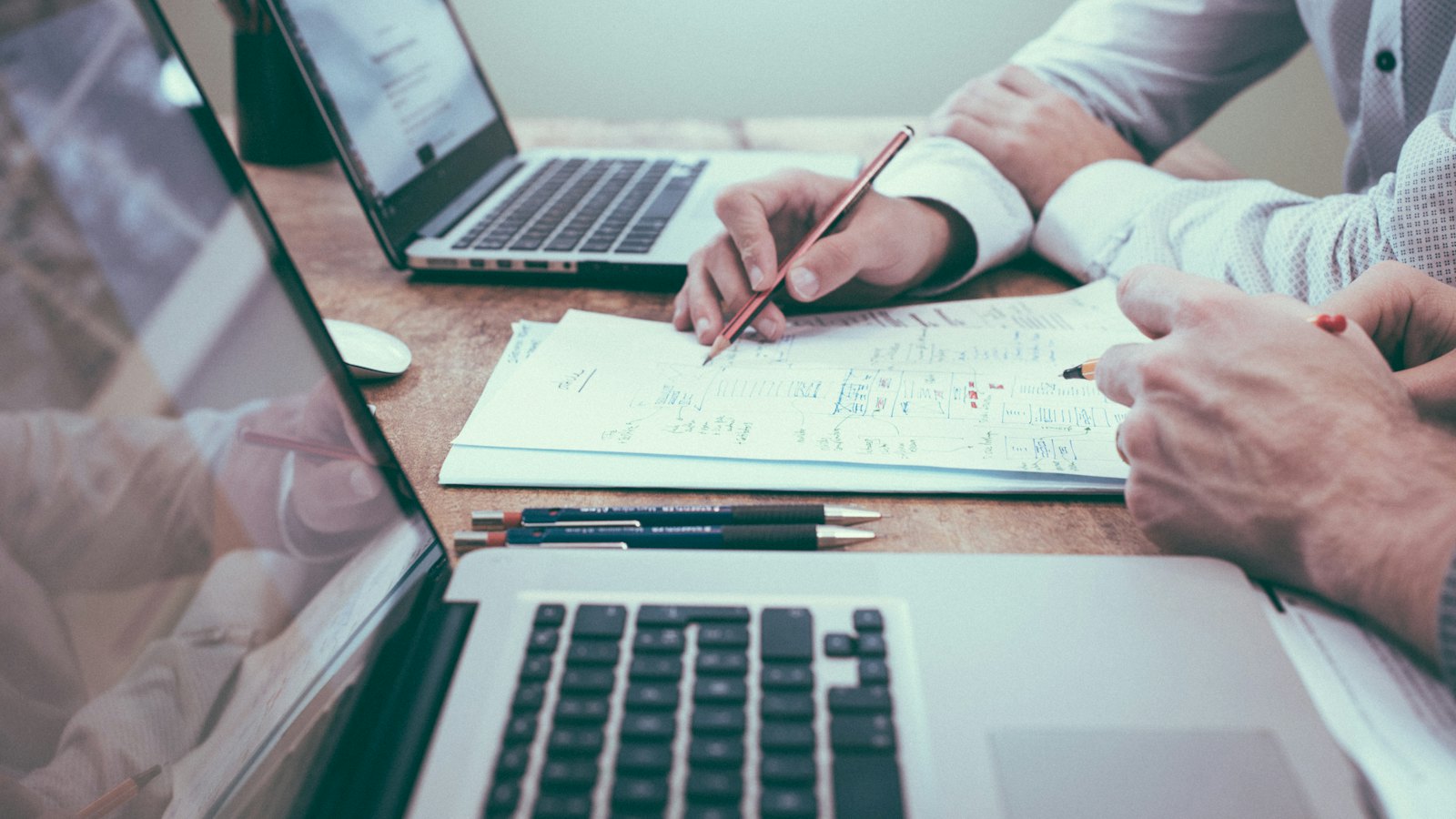 Duas pessoas revisando documentos em uma mesa com notebooks